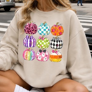 May include: A sand-colored crewneck sweatshirt featuring a graphic print of nine pumpkins in a grid layout. Each pumpkin displays a unique pattern, including leopard print, checkered, striped, and ghost motifs, accented with colorful bows.