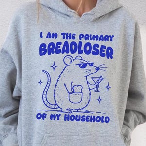 May include: A sport grey hooded sweatshirt featuring a blue graphic print of a rat wearing sunglasses, holding a martini glass, and standing with a slice of bread in its pocket. The text reads I AM THE PRIMARY BREADLOSER OF MY HOUSEHOLD.
