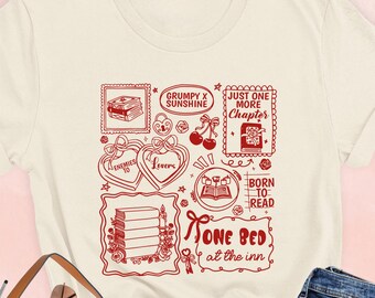 May include: Cream-colored t-shirt with a red graphic featuring book-themed illustrations and text. The text includes phrases like "Grumpy x Sunshine," "Just One More Chapter," and "Born to Read." The shirt is paired with blue jeans and a brown handbag.