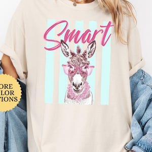 May include: Ivory-colored t-shirt featuring a graphic of a donkey wearing pink glasses and a floral collar, with the word "Smart" in pink script. The design is set against a backdrop of light blue vertical stripes. The shirt has rolled-up sleeves.