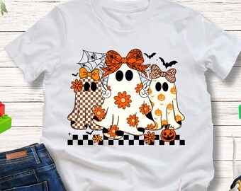 May include: White t-shirt featuring a Halloween-themed graphic of three ghosts with orange bows and flower details. The ghosts have different patterns, including checkered and floral designs. The shirt is paired with blue jeans and colorful sneakers.