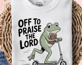 May include: White sweatshirt with the text "OFF TO PRAISE THE LORD" and a cartoon frog riding a scooter while holding a book. The sweatshirt is folded on a woven basket, with a cup of coffee and jeans in the background.
