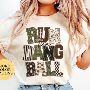 May include: An ivory short-sleeved t-shirt featuring a graphic print with the text RUN THE DANG BALL. The letters are filled with various football-themed patterns in shades of green, brown, and cream, including helmets, laces, and stripes.