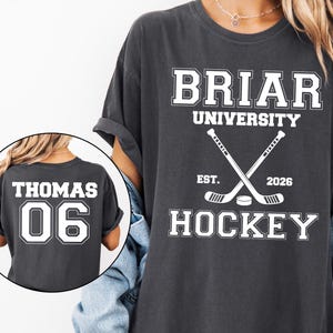 May include: A charcoal gray t-shirt featuring white collegiate-style text that reads BRIAR UNIVERSITY HOCKEY EST. 2026 with crossed hockey sticks and a puck. An inset circle shows the back of the shirt with the name THOMAS and the number 06.