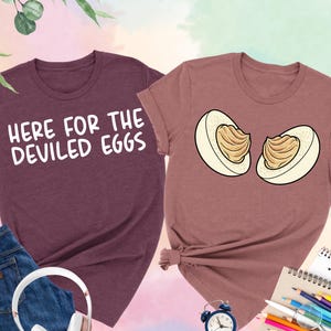 May include: Two heather t-shirts, one maroon and one mauve, with egg-themed graphics. One shirt says "HERE FOR THE DEVILED EGGS" in white text. The other shirt features a cartoon image of two deviled egg halves. Accessories include jeans and headphones.