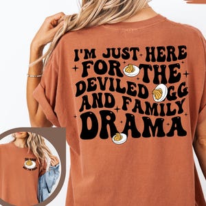 May include: A yam-colored t-shirt with the text "I'M JUST HERE FOR THE DEVILED EGG AND FAMILY DRAMA" in black. The design includes illustrations of deviled eggs. The shirt has short sleeves and a relaxed fit.