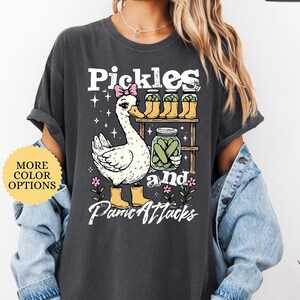 May include: Dark gray t-shirt with a whimsical graphic featuring a goose wearing a pink bow and yellow boots, alongside jars of pickles. The text reads "Pickles and Panic Attacks". A yellow circle says "More Color Options".