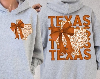 May include: A light gray hoodie featuring a Texas-shaped design with a floral pattern and a large orange bow. The back of the hoodie displays the word "TEXAS" in orange block letters. The hoodie is made of a soft material.