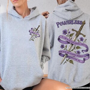 Puede incluir: Sudadera con capucha gris deportivo con bolsillo delantero y capucha. La parte delantera presenta una espada y un dise&ntilde;o floral con una cinta morada. La parte trasera muestra una espada, flores y el texto "POWERLESS" y una cita.
