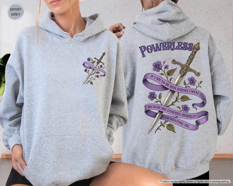 Puede incluir: Sudadera con capucha gris deportivo con bolsillo delantero y capucha. La parte delantera presenta una espada y un dise&ntilde;o floral con una cinta morada. La parte trasera muestra una espada, flores y el texto "POWERLESS" y una cita.