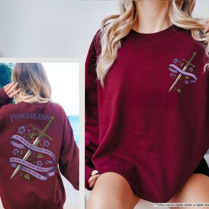 Puede incluir: Sudadera granate con un dise&ntilde;o de espada y flores en la parte delantera y trasera. La parte trasera de la sudadera tiene la palabra "POWERLESS" encima de una espada y una pancarta con texto. La parte delantera tiene un dise&ntilde;o de espada y flores m&aacute;s peque&ntilde;o.