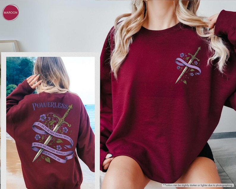 Puede incluir: Sudadera granate con un dise&ntilde;o de espada y flores en la parte delantera y trasera. La parte trasera de la sudadera tiene la palabra "POWERLESS" encima de una espada y una pancarta con texto. La parte delantera tiene un dise&ntilde;o de espada y flores m&aacute;s peque&ntilde;o.