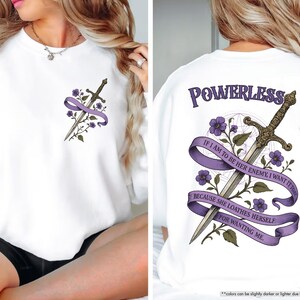 Puede incluir: Sudadera blanca con un dise&ntilde;o de espada y detalles morados. La parte trasera muestra la palabra "POWERLESS" y el texto: "IF I AM TO BE HER ENEMY, I WANT IT TO... BECAUSE SHE LOATHES HERSELF FOR WANTING ME." Elementos florales completan el dise&ntilde;o.