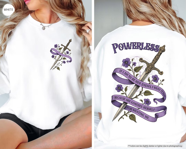 Puede incluir: Sudadera blanca con un dise&ntilde;o de espada y detalles morados. La parte trasera muestra la palabra "POWERLESS" y el texto: "IF I AM TO BE HER ENEMY, I WANT IT TO... BECAUSE SHE LOATHES HERSELF FOR WANTING ME." Elementos florales completan el dise&ntilde;o.