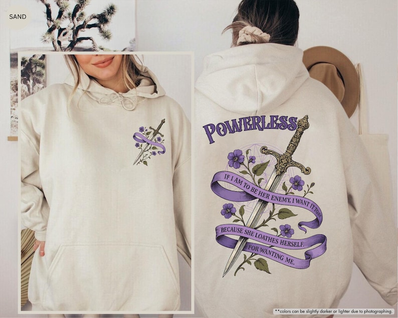 Puede incluir: Sudadera con capucha color arena con bolsillo delantero y capucha con cord&oacute;n. La parte delantera presenta una cinta morada, una espada y un dise&ntilde;o floral. La parte trasera muestra la palabra "POWERLESS" sobre una espada y una pancarta con texto.