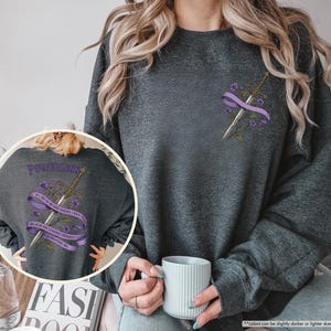 Puede incluir: Sudadera gris oscuro con un dise&ntilde;o de espada y flores. El dise&ntilde;o incluye una espada, una cinta morada y flores. La parte trasera de la sudadera tiene la palabra "POWERLESS" y el texto "IF I AM TO BE HER ENEMY I MUST BECOME HER BECAUSE SHE LOOKED HERSELF FOR WANTING ME."