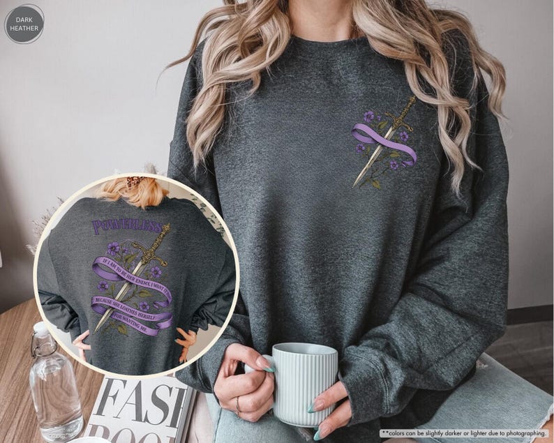 Puede incluir: Sudadera gris oscuro con un dise&ntilde;o de espada y flores. El dise&ntilde;o incluye una espada, una cinta morada y flores. La parte trasera de la sudadera tiene la palabra "POWERLESS" y el texto "IF I AM TO BE HER ENEMY I MUST BECOME HER BECAUSE SHE LOOKED HERSELF FOR WANTING ME."