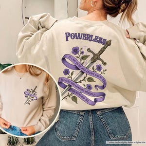 Puede incluir: Una sudadera de color arena con un dise&ntilde;o gr&aacute;fico que presenta una espada, flores moradas y el texto "POWERLESS" y una cita. El dise&ntilde;o est&aacute; en la espalda y una versi&oacute;n m&aacute;s peque&ntilde;a en la parte delantera.