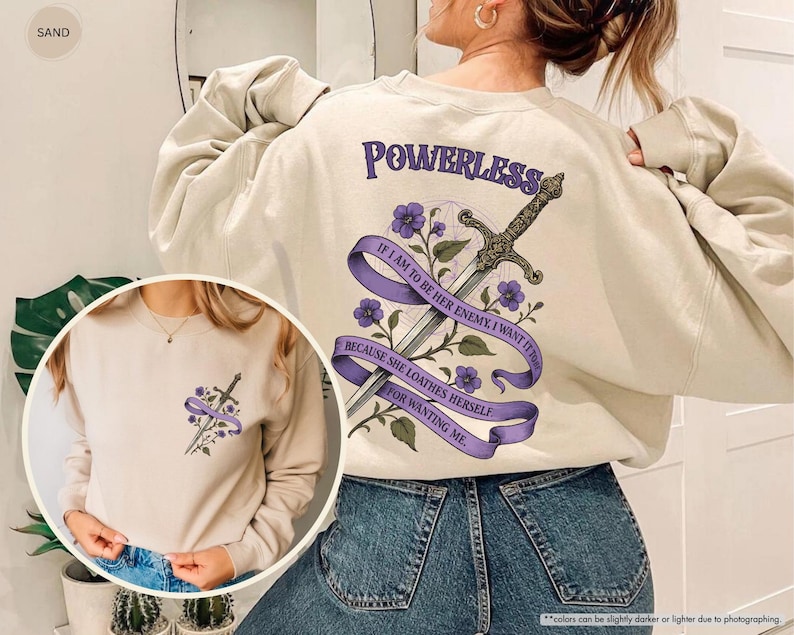 Puede incluir: Una sudadera de color arena con un dise&ntilde;o gr&aacute;fico que presenta una espada, flores moradas y el texto "POWERLESS" y una cita. El dise&ntilde;o est&aacute; en la espalda y una versi&oacute;n m&aacute;s peque&ntilde;a en la parte delantera.