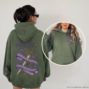 Puede incluir: Una sudadera con capucha verde oliva con la palabra "POWERLESS" sobre una espada y un dise&ntilde;o floral. El texto de la sudadera con capucha dice: "IF I AM TO BE HER ENEMY I WANT IT TO BE BECAUSE SHE LOATHES HERSELF FOR WANTING ME."