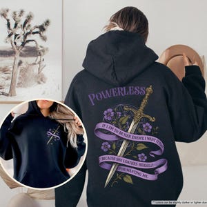 Puede incluir: Sudadera con capucha negra con un dise&ntilde;o gr&aacute;fico en la espalda. El dise&ntilde;o incluye una espada, flores moradas y el texto "POWERLESS" y una cita. La sudadera tiene un bolsillo delantero y una capucha.