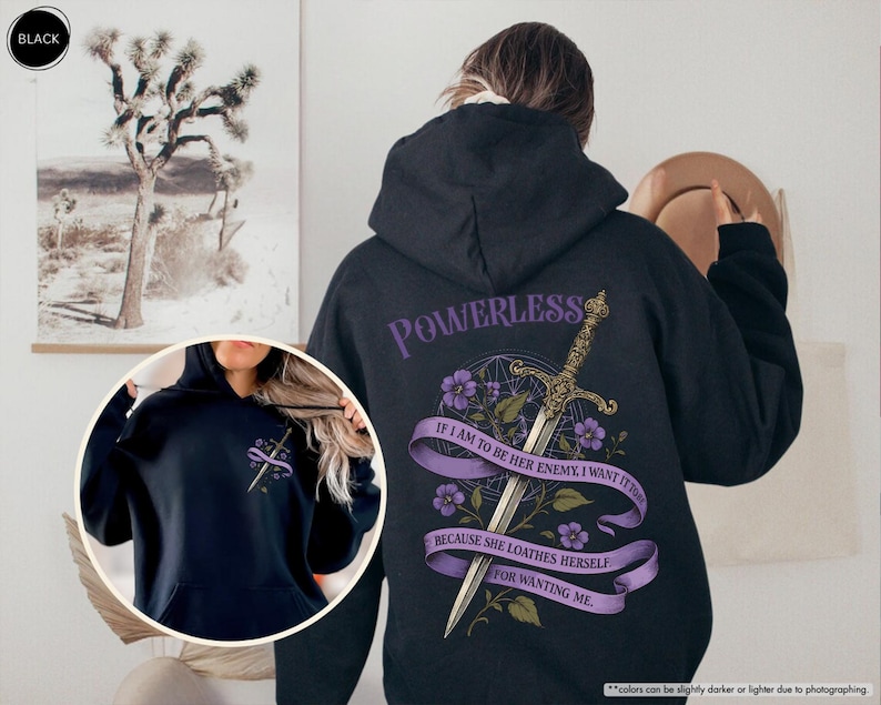 Puede incluir: Sudadera con capucha negra con un dise&ntilde;o gr&aacute;fico en la espalda. El dise&ntilde;o incluye una espada, flores moradas y el texto "POWERLESS" y una cita. La sudadera tiene un bolsillo delantero y una capucha.