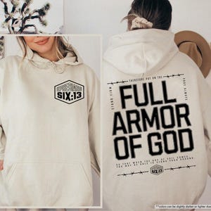 May include: Sand-colored hoodie with a front pocket and a hood. The front features a black and white hexagon graphic with the text "EPHESIANS SIX:13". The back displays "FULL ARMOR OF GOD" in bold black letters with additional text.