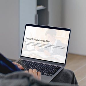 May include: A person using a laptop with the text "SAT/ACT Vocabulary Builder" on the screen. The laptop is open, displaying text and an image of a student. The keyboard is visible, and the person's hands are on the keyboard.