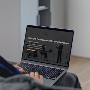 May include: A person using a laptop displaying a "Training & Development Planning Template." The screen shows a presentation with text and illustrations. The laptop is open, and the user's hands are visible on the keyboard.