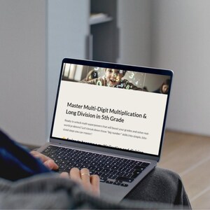 May include: A person using a laptop displaying a webpage titled "Master Multi-Digit Multiplication & Long Division in 5th Grade." The screen shows a child and numbers. The laptop is silver and has a black keyboard.