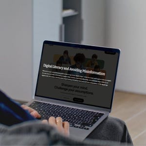 May include: A person using a silver laptop with the text "Digital Literacy and Avoiding Misinformation" on the screen. The laptop keyboard is visible, and the user's hands are on the keyboard. The screen also displays the text "Sharpen your mind. Challenge your assumptions."
