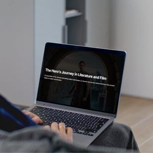 May include: A person using a silver laptop displaying the text "The Hero's Journey in Literature and Film." The screen shows an image of a man in a film scene. The laptop is on a person's lap.