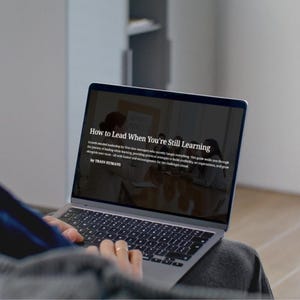 以下が含まれることがあります： 「How to Lead When You're Still Learning」というテキストが表示されたラップトップ。画面にはリーダーシップと成長に関するテキストが表示されています。ラップトップは開いており、「by TRAIN HUMANS」というテキストも表示されています。人がラップトップを使用しています。