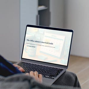May include: A laptop screen showing "The Office Admin's Survival Guide." The screen displays a calendar and a mug that says "World's Best Boss." The laptop is open on a person's lap, with their hands on the keyboard.