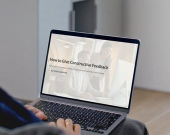 May include: A laptop displaying a presentation titled "How to Give Constructive Feedback." The screen shows a business setting with two people. The laptop is open, and the keyboard is visible. The text "by TRAIN HUMANS" is also visible.