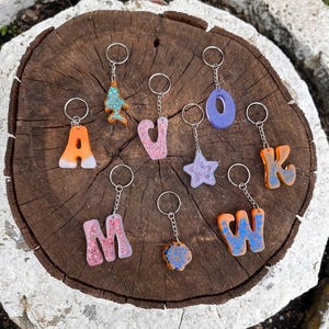 May include: A collection of 10 resin keychains featuring various letters, a star, and a Christmas tree. The keychains are made with translucent resin and have glitter embedded in them. The colors of the keychains are purple, orange, pink, and blue.