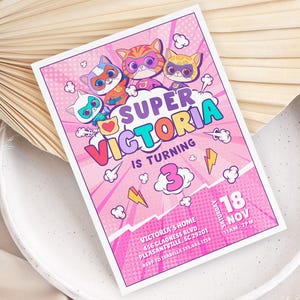 Puede incluir: Una invitación de cumpleaños rosa con el texto "Super Victoria cumple 3 años". La invitación presenta gatos superhéroes de dibujos animados e incluye detalles del evento: fecha, hora y lugar.
