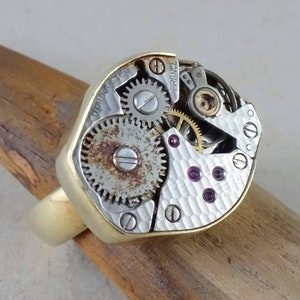 May include: A gold ring with a watch movement embedded in the top. The watch movement is silver and has a textured surface. The watch movement has several gears and a small red jewel. The watch movement is labeled "Jonvilisted France".