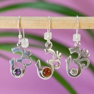 May include: Three silver-toned Om symbol earrings with different colored gemstones: purple, amber, and clear. The earrings hang from a wooden dowel against a soft purple background, with green leaves in the background.