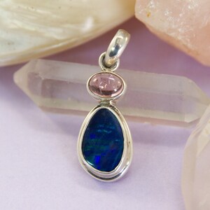 May include: A silver pendant with a blue opal gemstone and two pink gemstones. The pendant is on a white background.