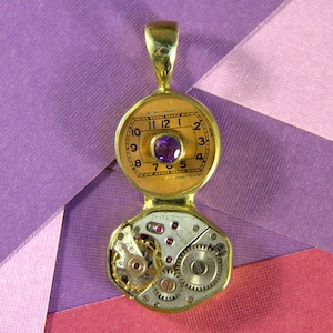 May include: A gold-toned pendant necklace with a round, open-faced pocket watch charm. The watch face has Roman numeral hour markers and a purple gemstone at the center. The back of the pendant reveals a detailed, intricate clockwork mechanism.