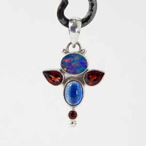 May include: A silver pendant featuring an oval, iridescent opal, two teardrop-shaped red garnets, a blue kyanite, and a small red garnet. The pendant is suspended from a metal loop. The gemstones are set in a silver setting.