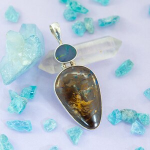May include: A silver pendant with a teardrop-shaped opal gemstone set in a silver bezel. The opal is a mix of brown, gold, and blue colors. The pendant is surrounded by small blue crystals.