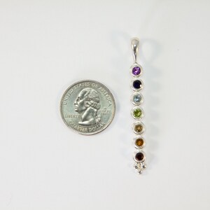 May include: A silver pendant with seven colorful gemstones in a vertical row. The gemstones are purple, dark blue, light blue, green, yellow, orange, and red. A U.S. quarter is next to the pendant for size comparison.