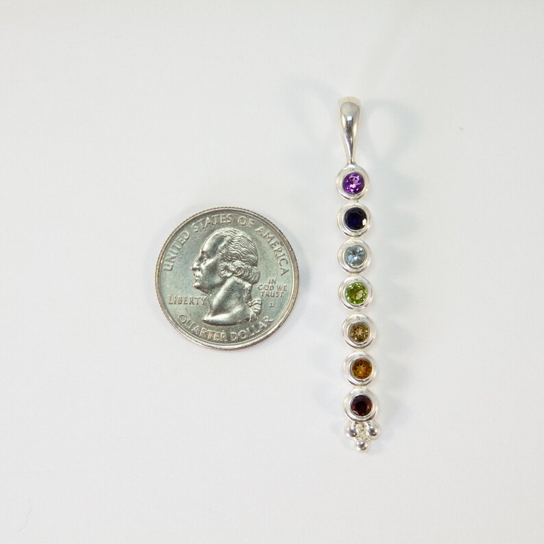 May include: A silver pendant with seven colorful gemstones in a vertical row. The gemstones are purple, dark blue, light blue, green, yellow, orange, and red. A U.S. quarter is next to the pendant for size comparison.
