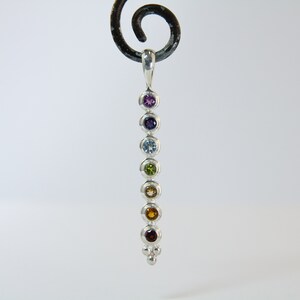 May include: A silver pendant featuring seven colorful gemstones in a vertical row. The gemstones include purple, indigo, light blue, green, yellow, orange, and red. The pendant has a small loop at the top and a cluster of three small silver beads at the bottom.
