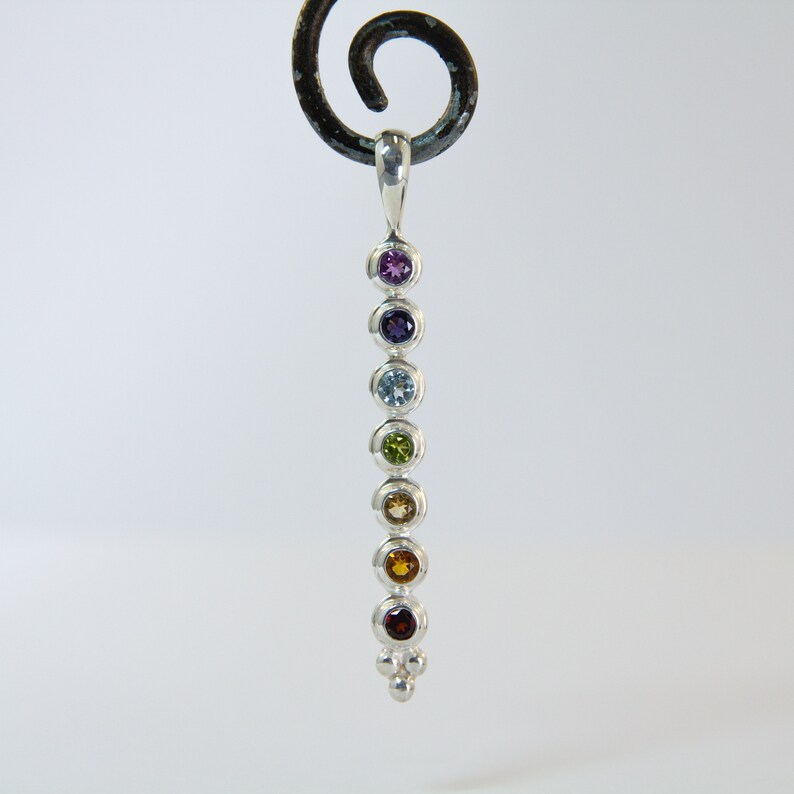 May include: A silver pendant featuring seven colorful gemstones in a vertical row. The gemstones include purple, indigo, light blue, green, yellow, orange, and red. The pendant has a small loop at the top and a cluster of three small silver beads at the bottom.