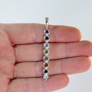 May include: A silver pendant featuring seven colorful gemstones in a vertical row. The gemstones are purple, indigo, light blue, green, yellow-brown, orange-brown, and red-brown. The pendant has a loop at the top for a chain and a decorative bottom.