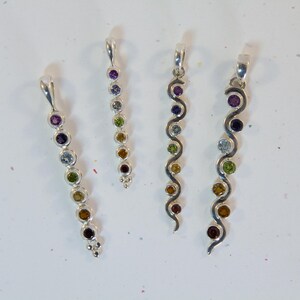 May include: Four silver pendants with colorful gemstones. The pendants have a loop at the top for a chain. The gemstones are various colors, including purple, blue, green, yellow, and brown. Two pendants have a straight design, and two have a wavy design.