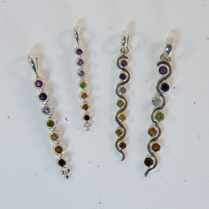 May include: Four silver pendants with colorful gemstones. The pendants have a loop at the top for a chain. The gemstones are various colors, including purple, blue, green, yellow, and brown. Two pendants have a straight design, and two have a wavy design.